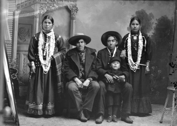 Full-length studio group portrait of two men, two Ho-Chunk woman, and a Ho-Chunk child. The men are sitting and wearing suits and hats, with the man on the right holding a boy standing in front of him. The women are standing and are wearing beads and earrings. Names (l to r) Unknown, John Rogue (CooseHeRaeKaw) (son of Young Rogue [WaChoWeKaw] and [WaKonHuGiWinKaw]) (from Winnebago, NE), Frank Washington Lincoln (Ho-Chunk) holding Charles Rogue, Henukaw Davis, Rogue. In the background is a painted backdrop.