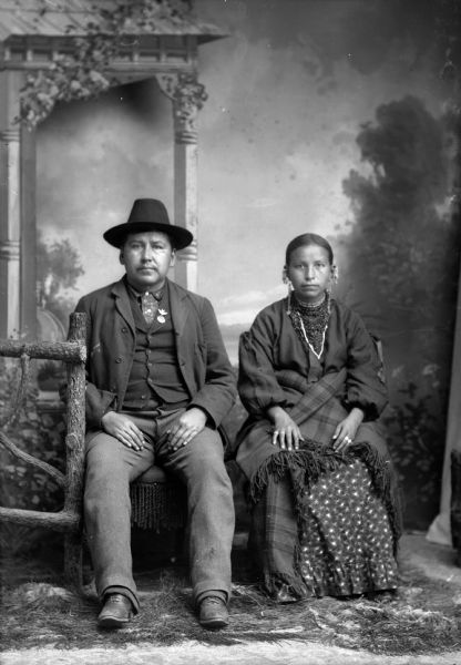Studio portrait of a Ho-Chunk man and woman, Sharpteeth Little Sam (he pa he ga se ba sik) and his wife, posing sitting in front of a painted backdrop. He is wearing a hat, suit jacket, vest with pin, (perhaps a military medal), and hat. She is wearing earrings, long skirt, and shawl around her waist.