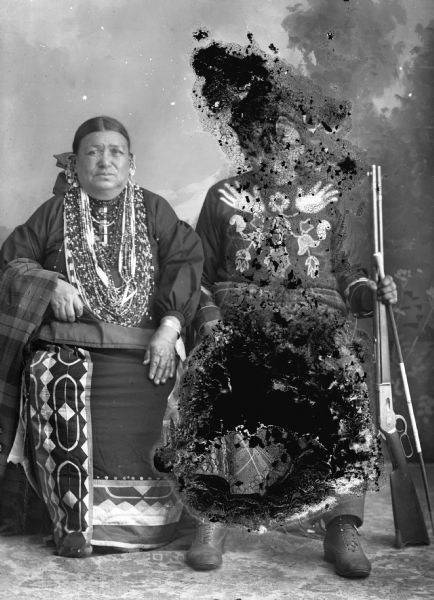 Studio portrait of a Ho-Chunk man and woman posing sitting and wearing regalia. The man is holding a rifle and another object upright with his left hand. Little Soldier (a.k.a., Strike the Tree, NoGinKah) and his wife, Bettie Littlesoldier (BayBayBawKah). They are posing in front of a painted backdrop.