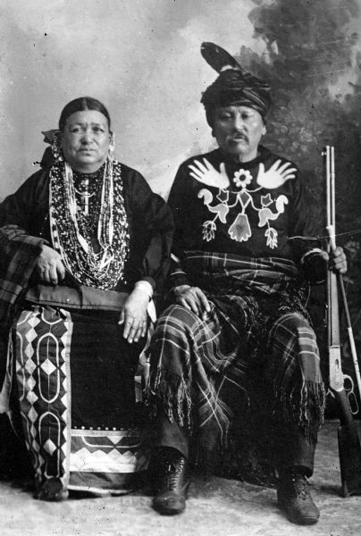 Copy photograph of a studio portrait of a Ho-Chunk man and woman posing sitting and wearing regalia, the former is also holding a rifle and another object in his left hand. Little Soldier (a.k.a., Strike the Tree, NoGinKah) and his wife, Bettie Littlesoldier (BayBayBawKah). They are in front of a painted backdrop.