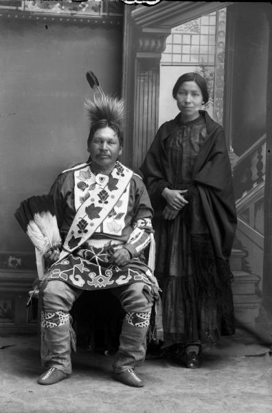 Studio portrait in front of a painted backdrop of a Ho-Chunk man posing sitting, with a Ho-Chunk woman in a shawl standing next to him. The man is wearing regalia, including Ho-Chunk beadwork. Alex Longtree and his daughter Emma Longtree Lowe, the wife of Gilbert Lowe.