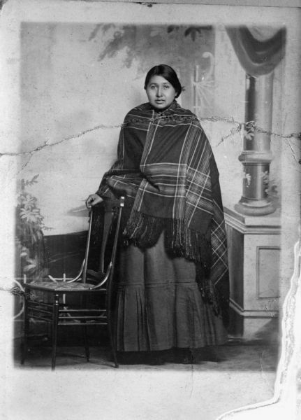 Copy photograph of a ripped print showing a Ho-Chunk woman, probably Lena Long Thunder, posing standing by a chair and wearing a plaid shawl in front of a painted backdrop.