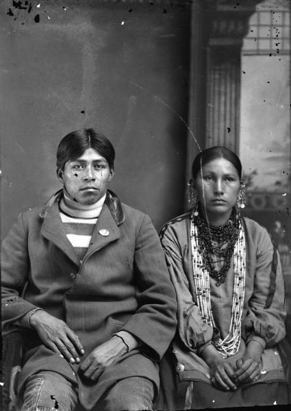 Studio portrait of a Ho-Chunk man and woman posing sitting in front of a painted backdrop. The man is wearing a coat and sweater. On the lapel is a pin with a portrait on it. The woman is wearing a blouse with ribbons, and beads and earrings. Possibly William Hall, Jr., and his wife, who was reportedly the sister of John Stacy.