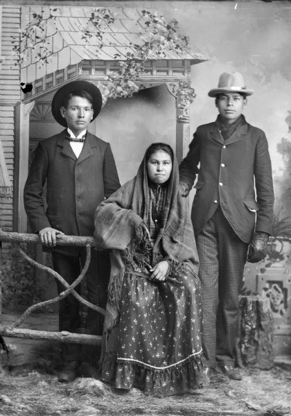 Studio portrait of a Ho-Chunk woman posing sitting on a prop wooden fence, and two Ho-Chunk men flanking her. The woman is wearing a shawl over her head, and the men are both wearing suit jackets and hats. The man on the left wearing the bow tie is Ed Mallory. They are posing in front of a painted backdrop.