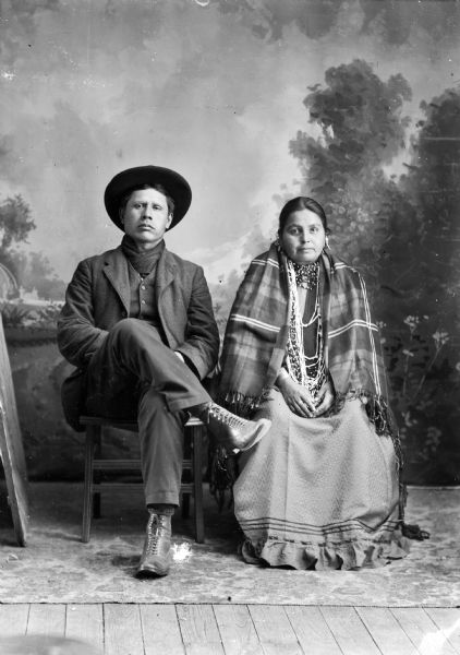 Studio portrait of a Ho-Chunk man, William Massey(Massie), and his wife "You Hear the Thunder," posing sitting in front of a painted backdrop. The former is wearing a suit jacket, vest, and hat, the latter is wearing beads and a wool shawl.