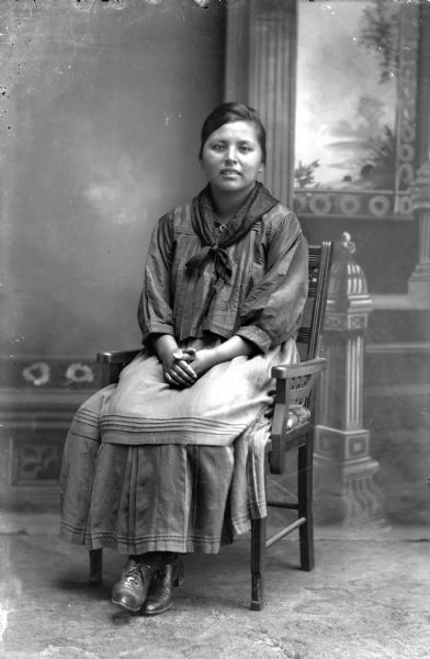 Studio portrait of a Ho-Chunk woman, Alice Redbird, posing sitting and wearing modern dress in front of a painted backdrop.