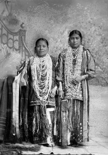 Studio portrait in front of a painted backdrop of two Ho-Chunk women posing standing. They are wearing dresses, and shell and bugle necklaces and silver ear bobs. The woman on the right is Red Bird Girl.