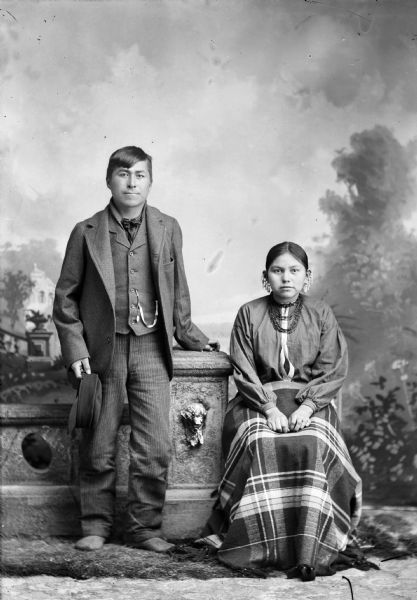 Studio portrait of a young Ho-Chunk man, Charles Smith, and his wife. The man is posing standing by a prop stone wall, and is wearing a suit jacket, vest with watch fob, and trousers, and she is wearing a wool shawl over her lap and ear bobs. They are posing in front of a painted backdrop.