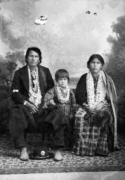 Studio portrait of a Ho-Chunk man, woman, and child. The couple is posing sitting and the child is posing standing between them. They are all wearing many beaded necklaces. Luke Snowball with his wife and child posing in front of a painted backdrop.