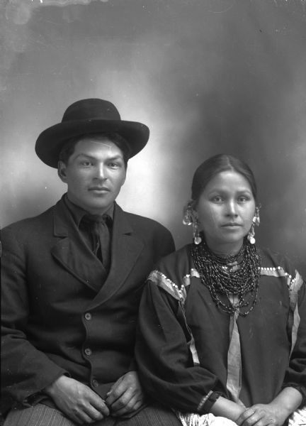 Studio portrait of a Ho-Chunk man and woman posing sitting in front of a painted backdrop. The man is wearing a suit, tie, and hat, and the woman is wearing necklaces, earrings, and a blouse decorated with ribbons. Henry Stacy and Mountain Wolf Woman, the youngest sister of Sam Blowsnake.