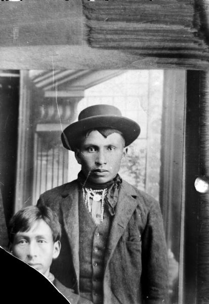 Copy photograph of studio portrait of two Ho-Chunk men in front of a painted backdrop. The one on the left is obscured by negative damage and the other, John Stacy, is wearing a suit, bead necklace, and a hat.