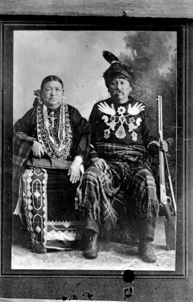 Copy photograph of a studio portrait of a Ho-Chunk man and woman posing sitting and wearing regalia. The man is also holding a rifle and another object in his left hand. Little Soldier (a.k.a., Strike the Tree, NoGinKah) and his wife, Bettie Littlesoldier (BayBayBawKah). They are posing in front of a painted backdrop.