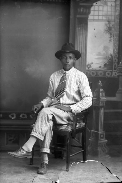 Studio portrait of a Ho-Chunk man, Luke Snowball Jr., posing sitting and wearing a shirt, tie, and hat in front of a painted backdrop.