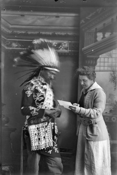 Studio portrait in front of a painted backdrop of a Ho-Chunk man in full regalia, including a Sioux headdress and holding a pipe, posing standing with a white woman in modern dress, who is looking at a letter. Identified as Thomas Thunder and Rachel Commons or Francis Densmore.
