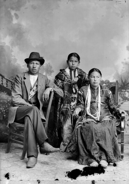 Studio portrait in front of a painted backdrop of a Ho-Chunk man, Thomas Thunder, posing sitting and wearing modern dress. A Ho-Chunk woman, his wife, is posing sitting and wearing several necklaces. Their child is posing standing between and slightly behind them, and is wearing necklaces and a shawl.