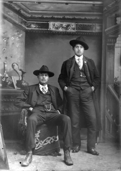 Studio portrait of two Ho-Chunk men, one posed standing and the other sitting, both wearing suit jackets, vests with watch fobs, ties, and hats. Probably John and Bill Thunder, posing in front of a painted backdrop.
