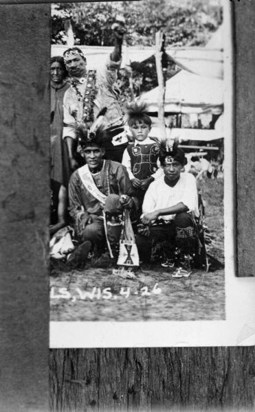 Copy photograph of image of Ho-Chunk men, two posing kneeling, with two people standing behind them on the left. One of the men standing has his left arm raised up. A boy is standing between the two kneeling men. All are wearing full Ho-Chunk regalia. The man on the left kneeling is probably Harold Jones Funmaker, also known as Wanajiska (trans. Fastest Man in Race), and the man in the background is Thomas Thunder.