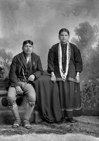 Studio portrait of a Ho-Chunk man, Henry Thunder, posing sitting and wearing modern dress along with a scarf, chain and claw necklace. A Ho-Chunk woman, his first wife, is posing standing and wearing several bead necklaces, file bracelets, and earrings. They are in front of a painted backdrop.