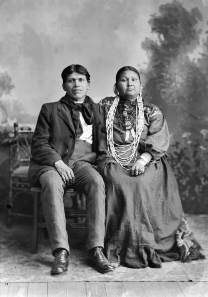 Studio portrait of a Ho-Chunk man and woman posing sitting in front of a painted backdrop. She is wearing nine strings of wampum and large ceramic trader beads. Henry Thunder and his second wife, Blowsnake Woman Thunder, the second daughter of Sam Blowsnake and mother of Alec Thunder.