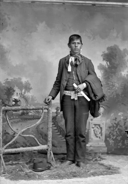Full-length studio portrait of Frank C. Thunder (Son of [WaConChaKah] John Thunder aka Dr. Thunder and [WeHonPeKaw] Lucy Bear, Thunder) standing next to a prop wooden fence. He is the brother of Thomas Thunder. He is wearing long earrings and a beaded sash, and holding what may be a rolled document in his hand and a wool shawl over his arm. There is a hat on the ground near the fence. In the background is a painted backdrop.