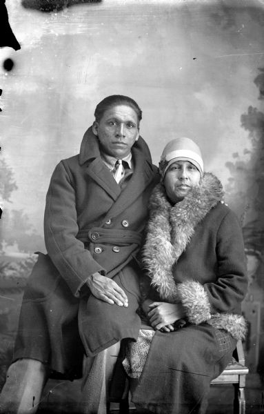 Studio portrait of a Ho-Chunk man posing sitting on the arm of a chair and leaning on a Ho-Chunk woman sitting in the chair. They are both wearing modern dress, including winter coats, and are in front of a painted backdrop. Frank Thunder and his wife, who was also married to St. Cyr.