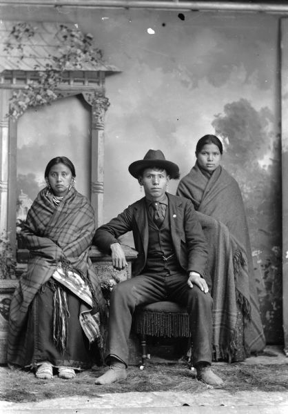 Studio portrait of a young Ho-Chunk man, Frank C. Thunder (son of [WaConChaKah] John Thunder aka Dr. Thunder and [WeHonPeKaw] Lucy Bear, Thunder), sitting, flanked by two young women. (l to r) Frank Thunder's wife, a Walkingcloud woman, and Kate Whitespirit-Dickson, Thunder, Decorra, Baptiste, Seymour (NeChooKoontchRaWinKah).  The two women are wrapped in shawls. In the background is a painted backdrop and prop stone wall.
