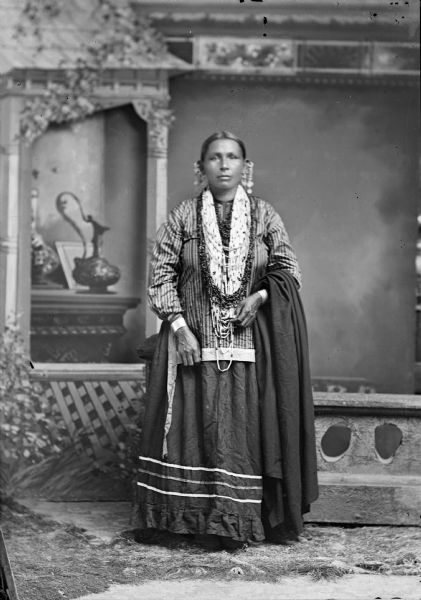 Studio portrait of a Ho-Chunk woman, Mary Thunderchief Snowball, posing standing in front of a painted backdrop near a prop stone wall. She is  holding a wool shawl and wearing several bead necklaces, file bracelets, and long coin earrings.