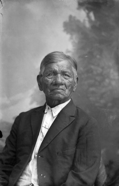 Studio portrait of an elderly Ho-Chunk man posing sitting in front of a painted backdrop. He is wearing a suit jacket and button-down shirt. Probably Moheeka Thundercloud.