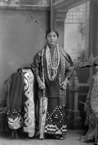 Studio portrait of a Ho-Chunk woman, Flora Thundercloud, posing standing in front of a painted backdrop. She is wearing several necklaces, a ribbon embroidered dress, and earrings, and is standing near a chair draped with an embroidered cloth.