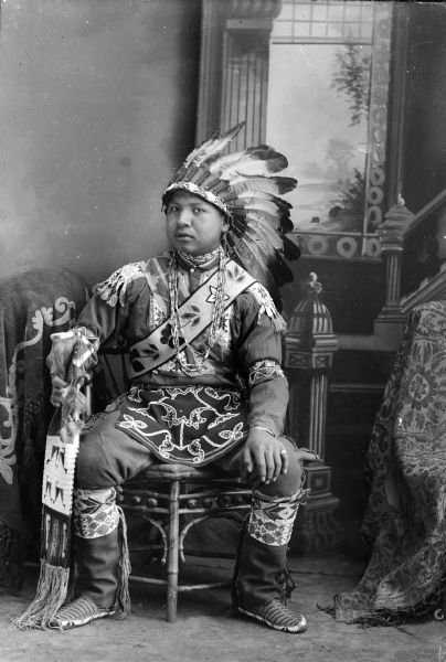 Studio portrait in front of a painted backdrop of a young Ho-Chunk man, Benjamin Thundercloud, posing sitting in regalia for initiation into the medicine lodge. He is holding a medicine pouch and is wearing a Sioux headdress, woodland flora breech cloth, beaded belt, Winnebago rings and bracelets, and moccasins.