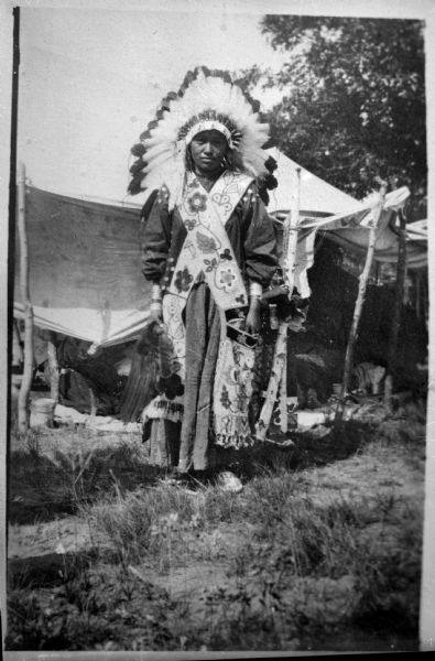 Copy photograph of a Ho-Chunk woman, standing outdoors in front of a tent, wearing a Sioux headdress and a Chippewa bandoleer.