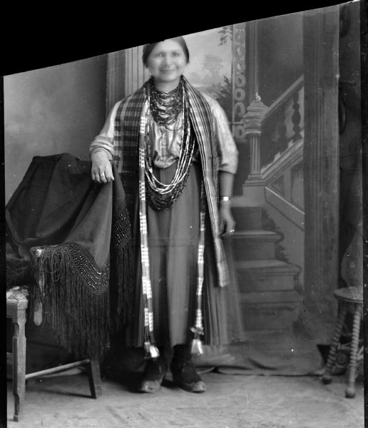 Studio portrait of a Ho-Chunk woman, Elizabeth Thundercloud Hall, posing standing near a chair draped with a four-inch row beaded and fringed shawl. She is wearing a dress and several necklaces in front of a painted backdrop.