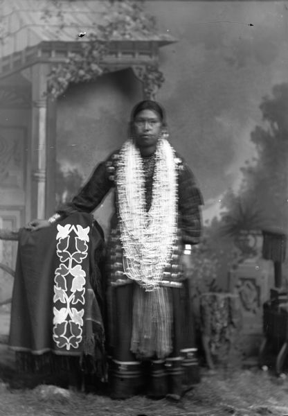 Studio portrait of a Ho-Chunk woman, Thunder Queen, posing standing near a chair in front of a painted backdrop. She is wearing many necklaces, and is displaying a woodland and flora ribbon-work shawl. She may have a goiter.