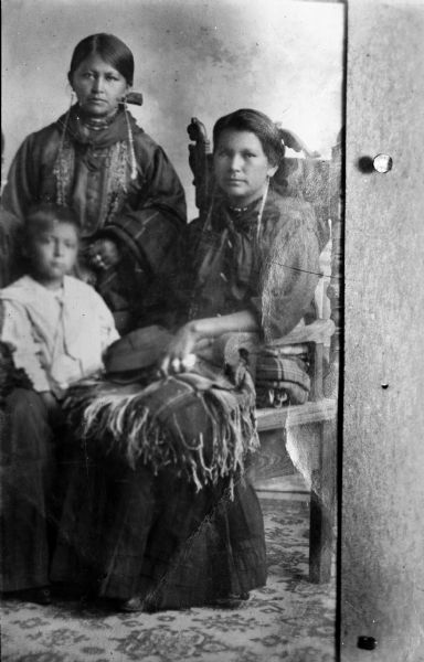 Copy photograph of a Ho-Chunk woman posing sitting and wearing long earrings and a shawl, and another woman and a child standing on the left. Clear Sky Winneshiek, the wife of Frank Winneshiek and mother of Sadie Winneshiek.