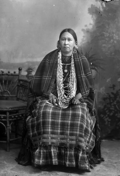Studio portrait of a Ho-Chunk woman posing sitting and wearing shawls, long necklaces, and chain and coin earrings. Wolf Woman, the sister of Alec Lonetree.