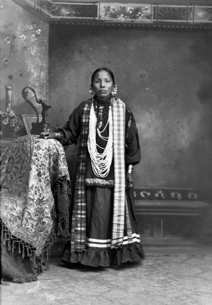 Studio portrait of a Ho-Chunk woman, posed standing with a hand resting on a stuffed chair in front of a painted backdrop. She is wearing coin and chain earrings, several long necklaces, and a plaid shawl over her shoulders.

