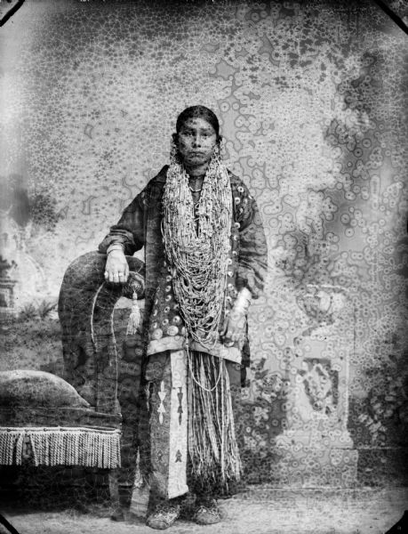 Studio portrait in front of a painted backdrop of a Ho-Chunk woman posing standing near a chair and wearing full regalia, including long necklaces, earrings, etc. The wife of George Otter and the oldest Youngswan daughter.