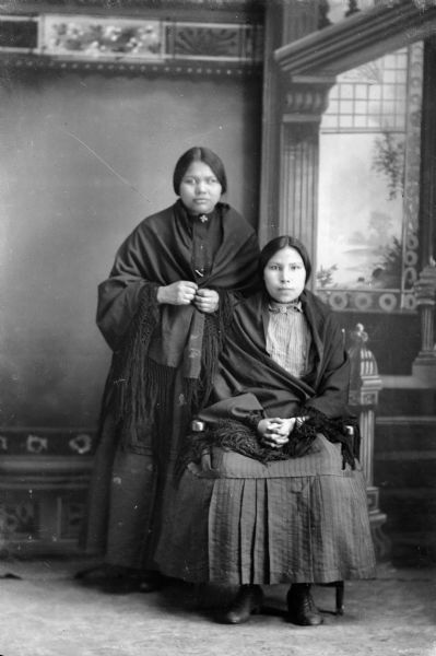 Studio portrait of two Ho-Chunk women posing in front of a painted backdrop. The woman on the left is standing and the woman on the right is sitting, and both are wearing contemporary dress and shawls. The woman on the right is ? Waukon.
