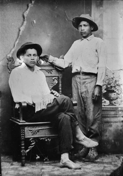 Copy photograph of a studio portrait of two Ho-Chunk men. The man on the right is standing, and the man on the left is sitting. Both men are wearing a hat and contemporary dress, and are posing in front of a painted backdrop.