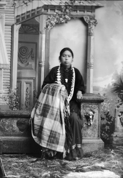 Studio portrait of a Ho-Chunk woman, Ethel/Esther White posing in front of a painted backdrop. She is sitting on a prop stone wall and is holding a plaid shawl, and is wearing bead necklaces, earrings, etc.