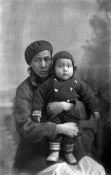 Studio portrait in front of a painted backdrop of a Ho-Chunk woman, Nancy Davis White Dog, sitting and holding a Ho-Chunk child. They are both dressed in contemporary winter clothing.