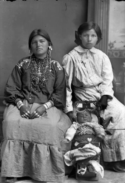 Studio portrait in front of a painted backdrop of two young Ho-Chunk women and a Ho-Chunk infant. The woman on the left is wearing regalia and the woman on the right is wearing contemporary clothing. The infant is in a papoose resting on the floor between them. The Windblowe sisters, on the left Nellie Windblowe Thundercloud, the wife of Adam Thundercloud, and her child, Koonoo. On the right Sarah Windblowe Decorah, wife of Rufus Decorah.