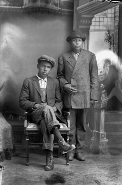 Studio portrait of two young Ho-Chunk men. Both men are wearing hats. The man sitting on the left, Simon Robert Littlesoldier (Wo No Ka Re Hunk Kah) (Son of David Bow Littlesoldier and Emma Thunder, Littlesoldier), is wearing a suit jacket and bib overalls. The man on the right, Art Decorah, is standing and wearing a double-breasted suit coat. In the background is a painted backdrop.
