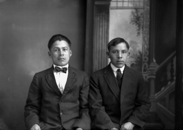 Studio portrait of two young Ho-Chunk men posing sitting and wearing suit jackets and ties in front of a painted backdrop. John White, on the left, is wearing a bow tie. On the right, Jesse Thompson (Little Bear), is wearing a necktie. He was reportedly killed in World War I and was the brother of John Thompson.