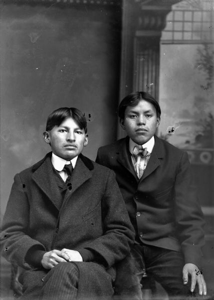 Studio portrait of two young Ho-Chunk men posing sitting in front of a painted backdrop. They are both wearing suit jackets. On the left, Black Coon of the Nebraska band is wearing a necktie. Pete Pettibone on the right is wearing a bow tie.