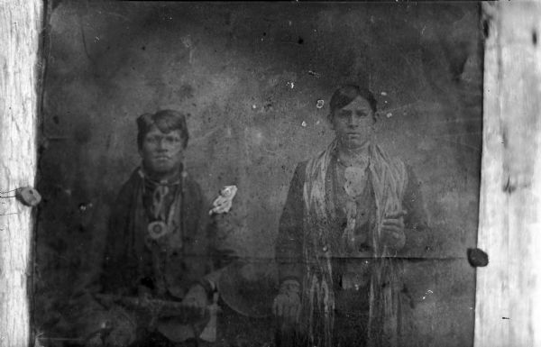 Copy photograph of a studio portrait of two young Ho-Chunk men posing standing and wearing regalia, including a finger-braided shawl on the man on the right. The man on the left is probably holding a metal tomahawk pipe. Identified as Henry Thunder on the left and Old George Funmaker, who is holding a cigar, on the right.