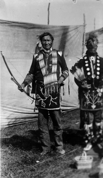 Copy photograph of a Ho-Chunk man, Jim Swallow, in regalia posing standing and holding a bow in front of cloth barriers or tents. From a powwow group from the 1908 Homecoming. Another man in regalia, William Massey(Massie), is out of focus on the right.