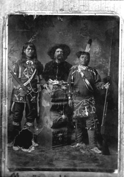 Copy photograph of a studio portrait of a European American man flanked by two Ho-Chunk men, all posing standing. The Ho-Chunk men are dressed in regalia and are holding rifles. Identified as Tom Roddy, with large moustache, standing in the center, and two members of his wild west show, any of the following: Crashing Thunder, Jasper Blowsnake, the father of Thomas Thunder, Alec Lonetree, or George Monegar.
