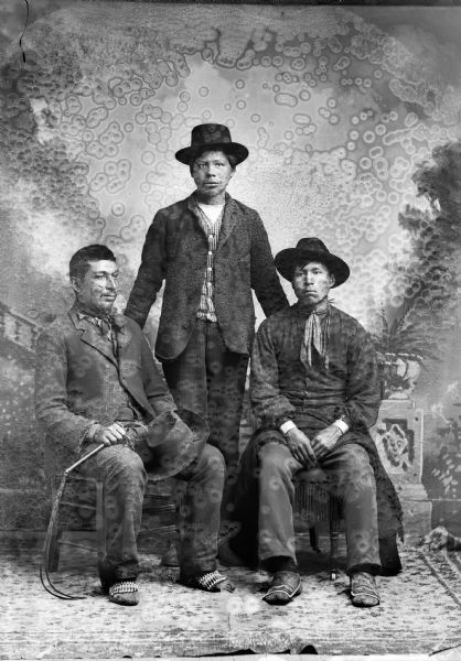 Studio portrait of (sitting, l to r) Joseph Thunderking and Jim Smith (Snowball) (Ma Ka Ko Jump Kah) in front of a painted backdrop. Standing in front is [????] Decorah. Joseph Thunderking is holding a hat and possibly a braided whip, the other men are wearing hats. Jim Smith is wearing a vest, and the other men are wearing suit jackets.