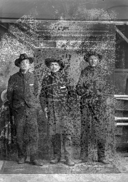 Studio portrait of three Ho-Chunk men posing standing in front of a painted backdrop. They are all wearing hats. The man on the left is wearing a suit jacket, the man in the center is wearing an overcoat, and the man on the right is wrapped in a blanket/shawl. Identified as Louis Johnson, Sr. in the center, and Howard McKee standing on the right.